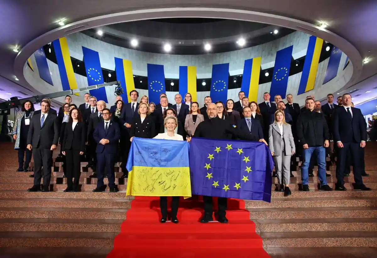 A formal group photo shows numerous officials standing on a red-carpeted staircase in a modern building. In the foreground…