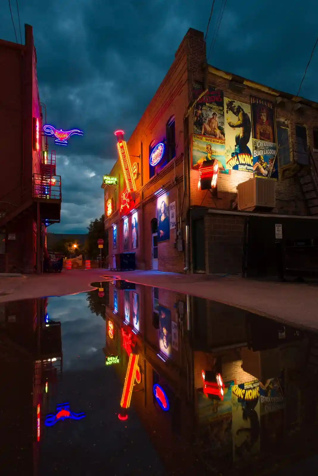 A nighttime photo of a brick alleyway with vintage neon signs and movie posters reflecting in a puddle. Visible signs…