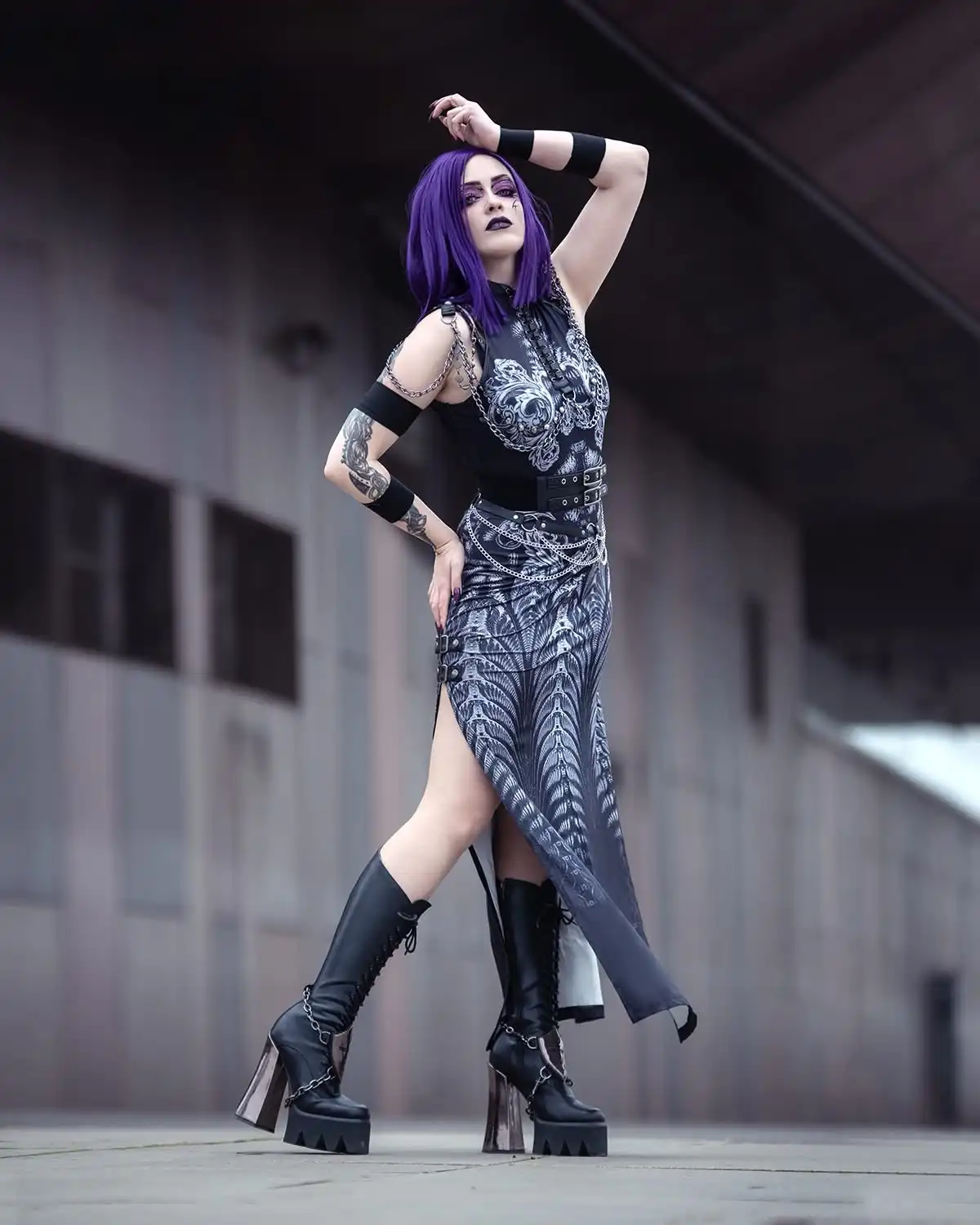 A person with purple hair and dark makeup poses confidently in a black and silver gothic outfit with chains, belts, and tall…