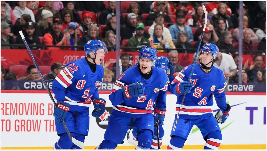 Three USA ice hockey players in blue uniforms celebrate enthusiastically on the ice, mouths open in excitement, with a crowd…