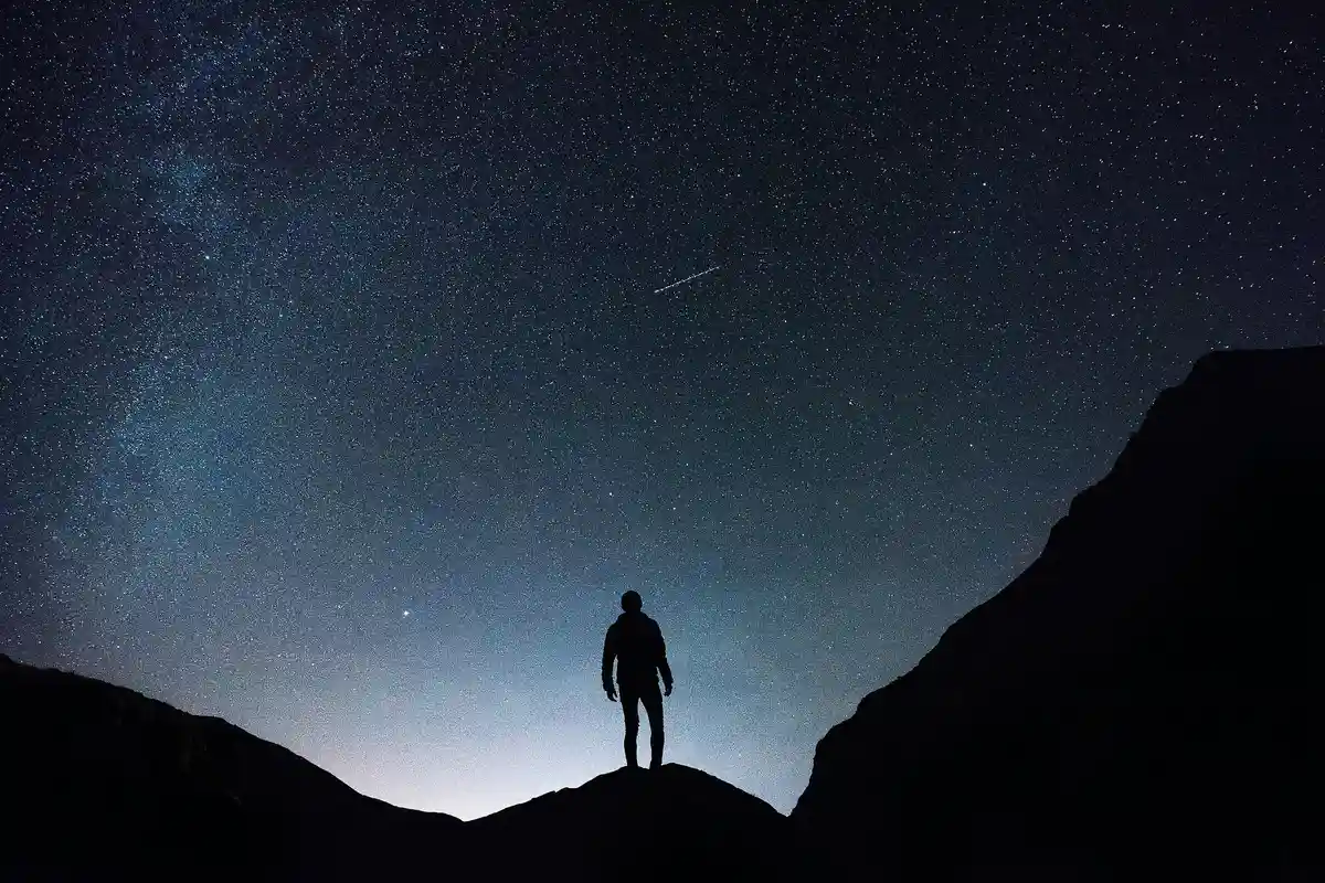 A silhouette of a person standing alone on a rocky peak under a vast, star-filled night sky with visible Milky Way and a…