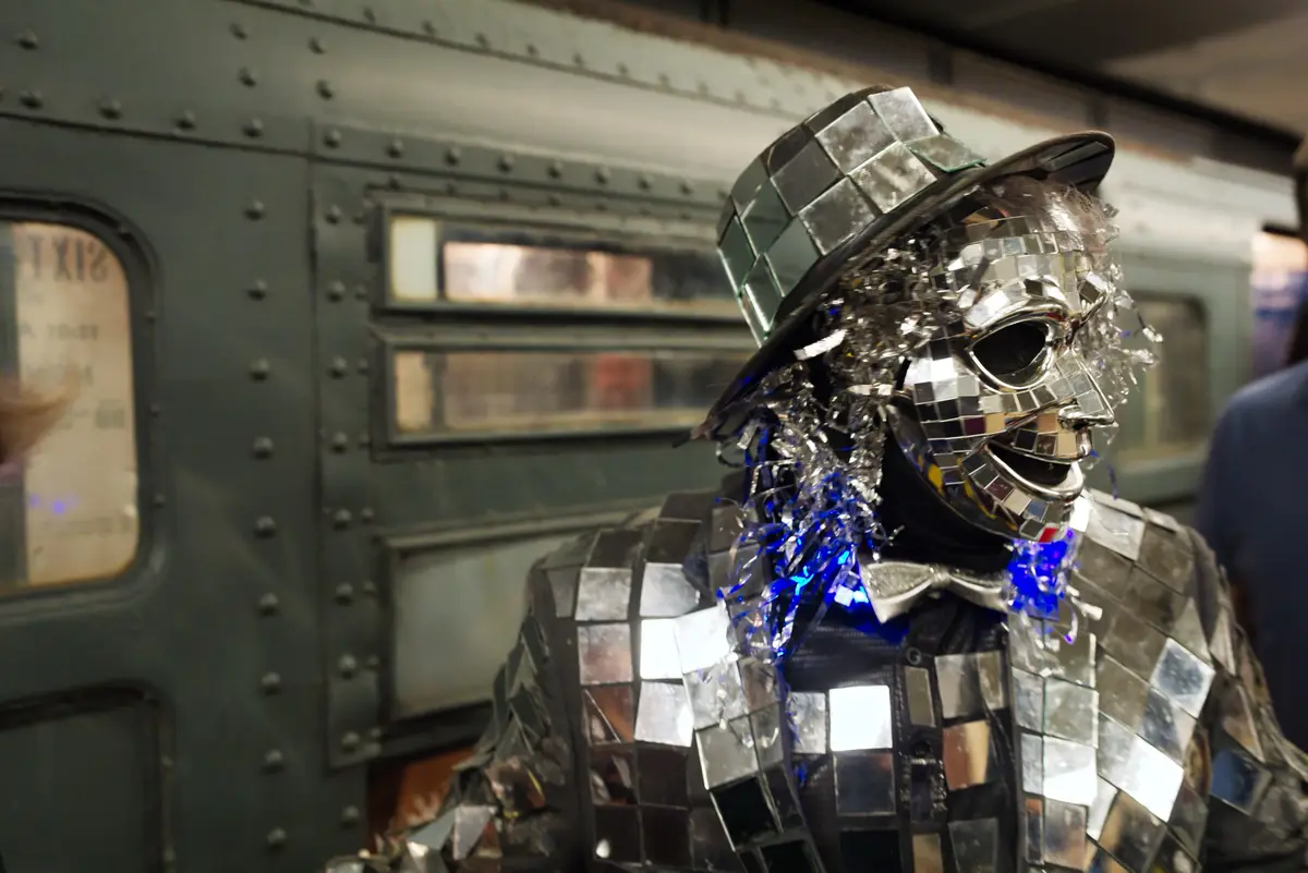 A person in a subway station is dressed in a full-body costume made of small, reflective mirror tiles, covering their face…