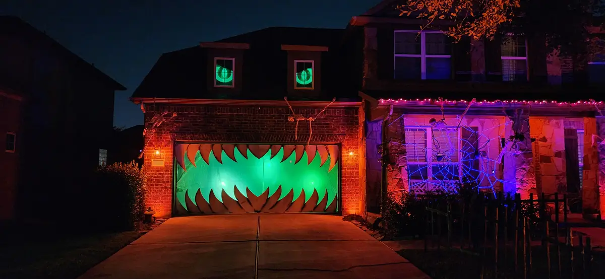 A house at night is decorated for Halloween with a large green glowing mouth on the garage door, glowing green eyes in the…