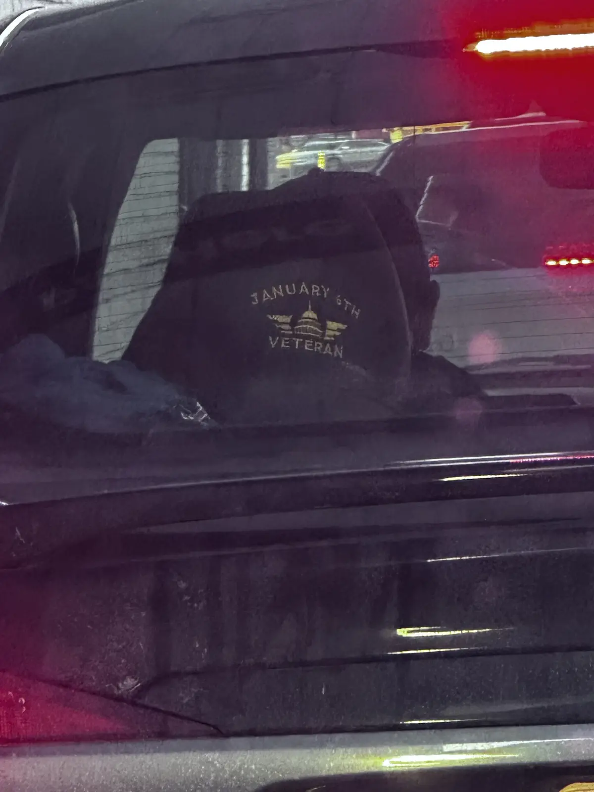 A person is seen from behind inside a car, wearing a black cap with gold lettering that reads 'JANUARY 6TH VETERAN' above a…