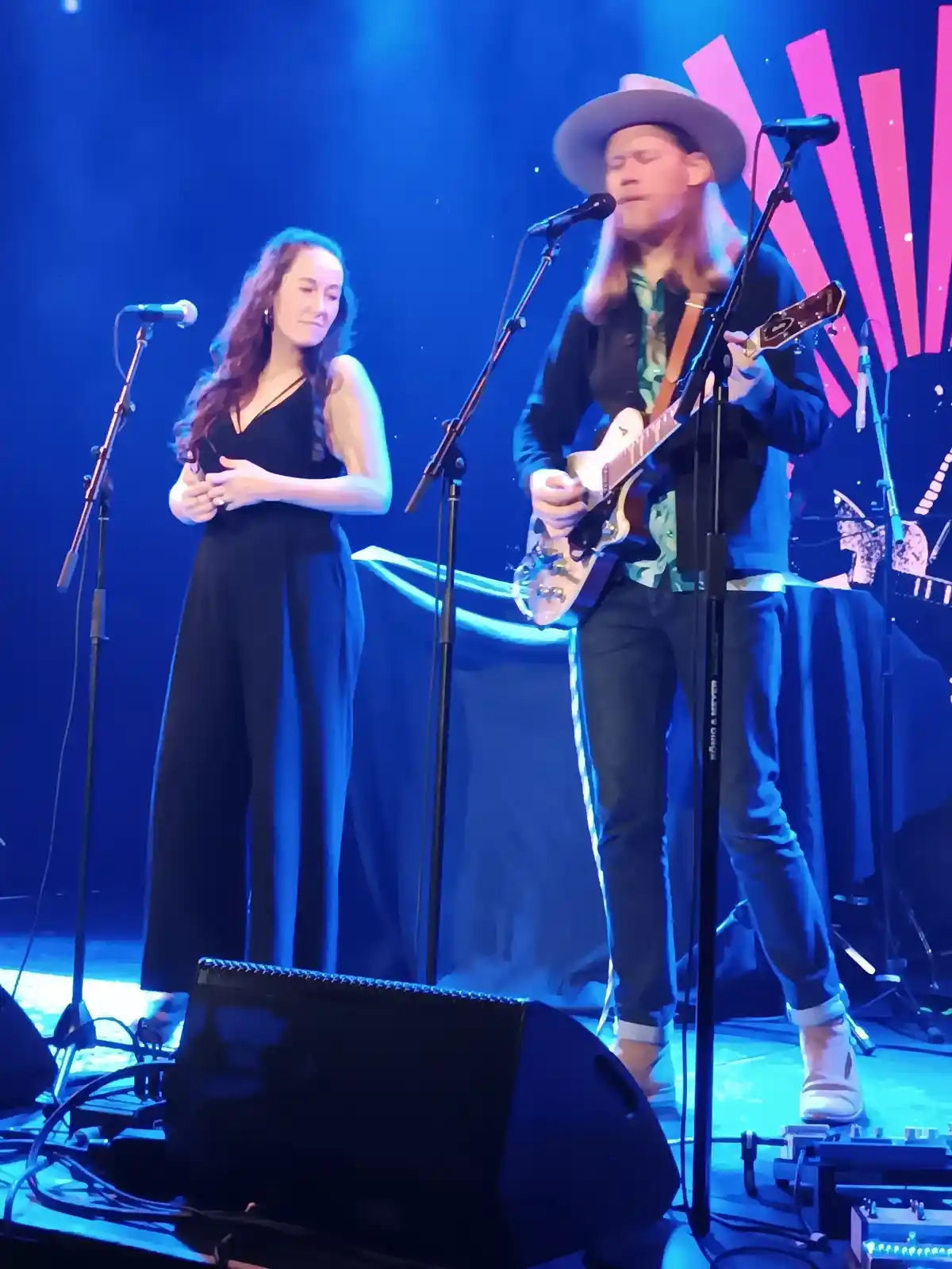 A man with long hair and a hat plays guitar on stage while a woman in a black jumpsuit stands beside him, both under blue…