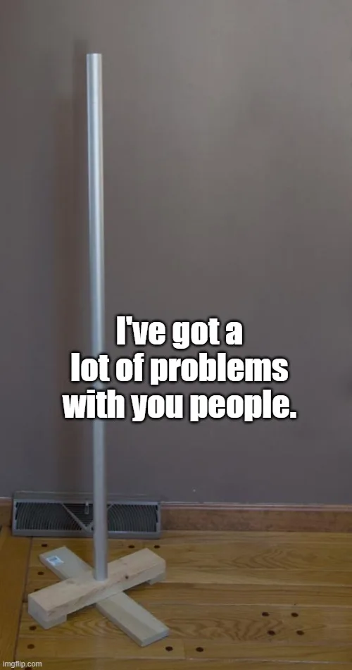 A tall, silver pole stands upright on a simple wooden crossbase on a hardwood floor, positioned in front of a plain brown…