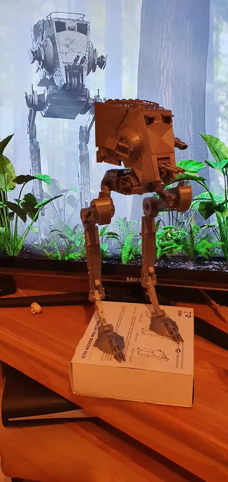 A brown AT-ST walker toy stands on a white product box on a wooden desk, with a TV screen in the background displaying a…