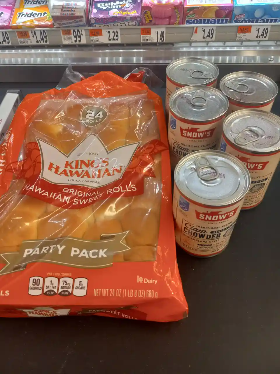 A grocery store counter displays a large orange bag of Kings Hawaiian Original Hawaiian Sweet Rolls Party Pack next to five…
