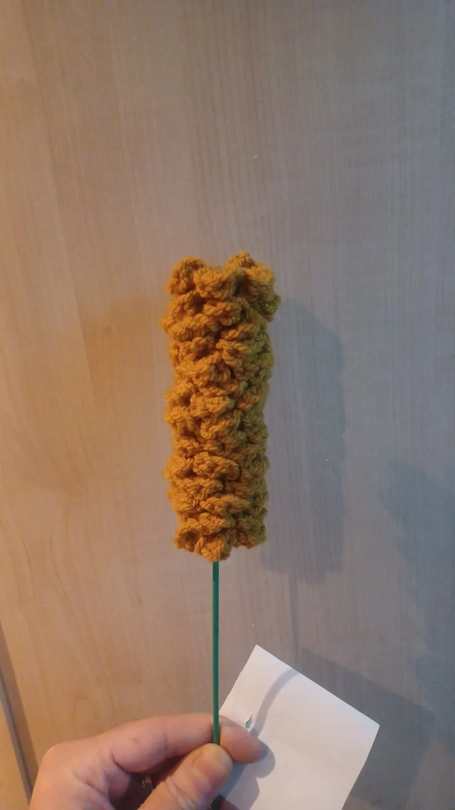 A hand holds a green stick with a crocheted, textured, golden-brown object resembling a corn dog on top, against a wooden…