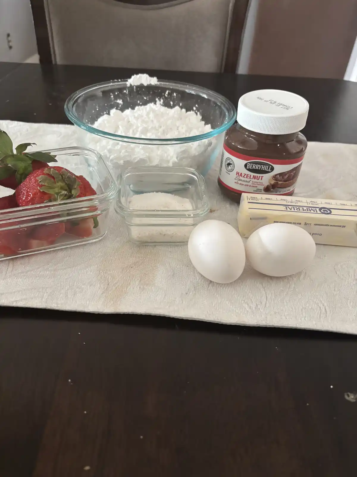 A photo of baking ingredients on a table: flour in a glass bowl, strawberries in a container, sugar, two eggs, butter, and a…