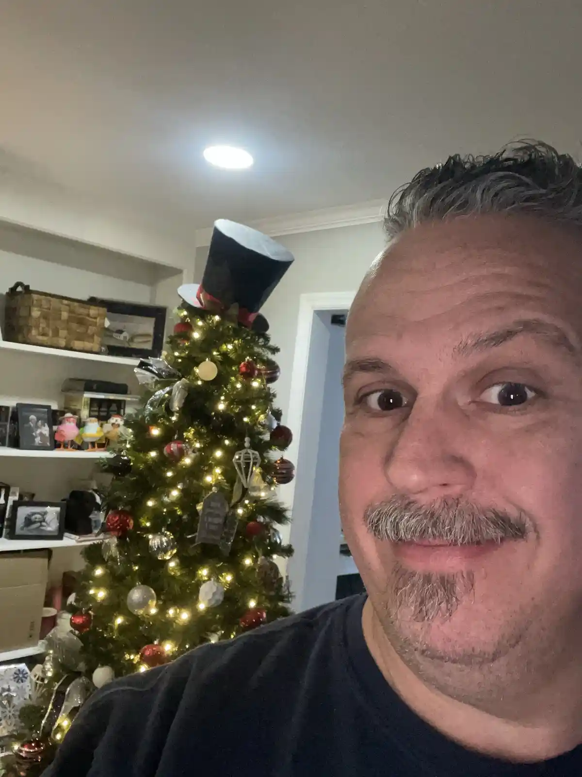 A man with a mustache and goatee takes a selfie in front of a decorated Christmas tree. The tree has lights, ornaments, and…