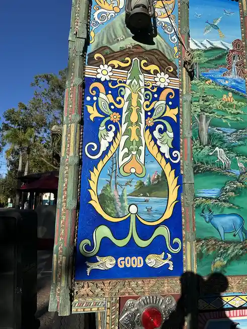 A colorful, ornate painted panel features a central blue oval with a pastoral scene of water, trees, and animals, framed by…