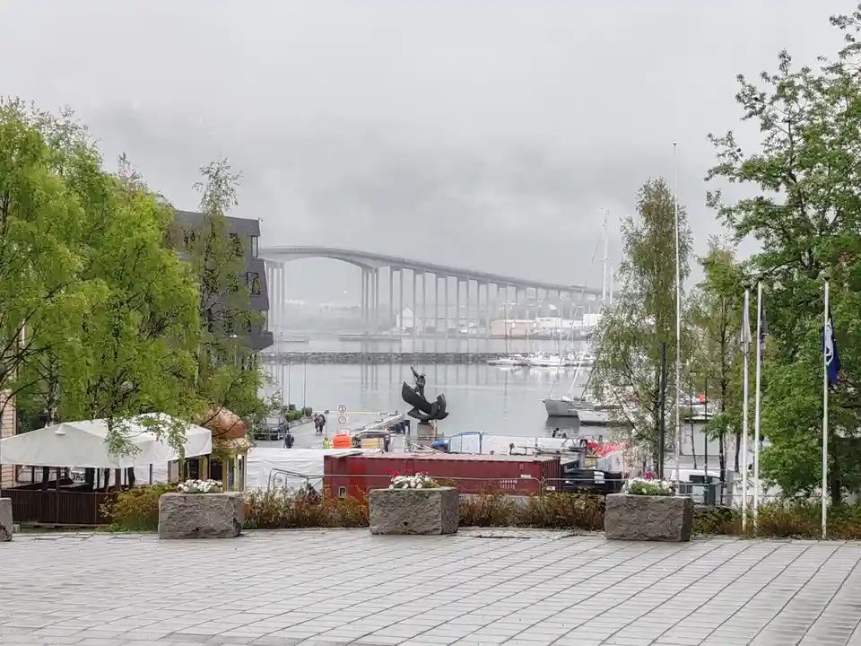 A gray, overcast day at a waterfront plaza with a large bridge spanning the water in the distance. Several sailboats are…