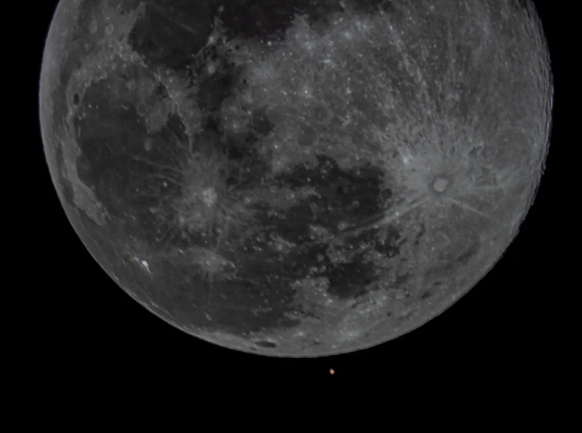 A close-up grayscale photograph of the moon's cratered surface against a black sky, with a tiny orange dot visible near the…