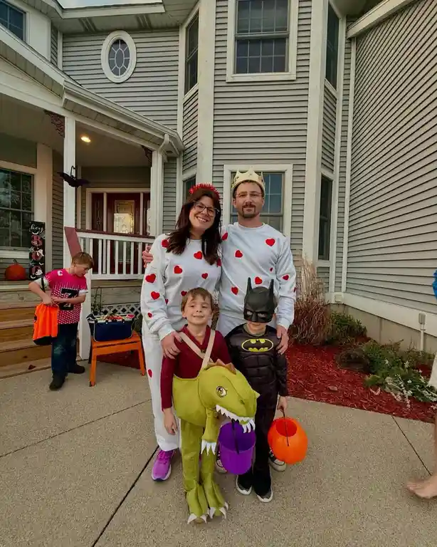 A family of four poses for a Halloween photo on their front porch, dressed in costumes. The parents wear matching white…