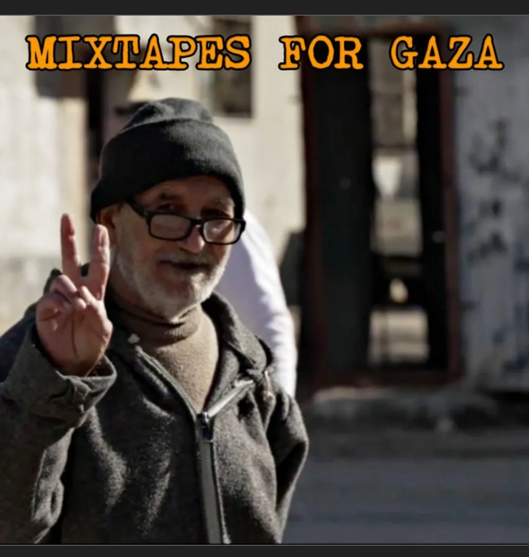 A man with glasses and a black beanie smiles while holding up a peace sign with his right hand. He wears a dark zip-up…