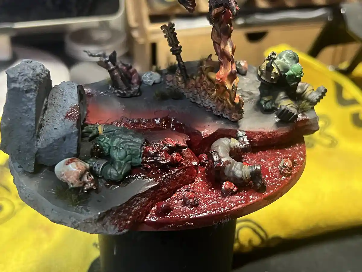 A highly detailed, gory miniature diorama depicts a battle scene with green-skinned figures, one lying dead in a pool of red…