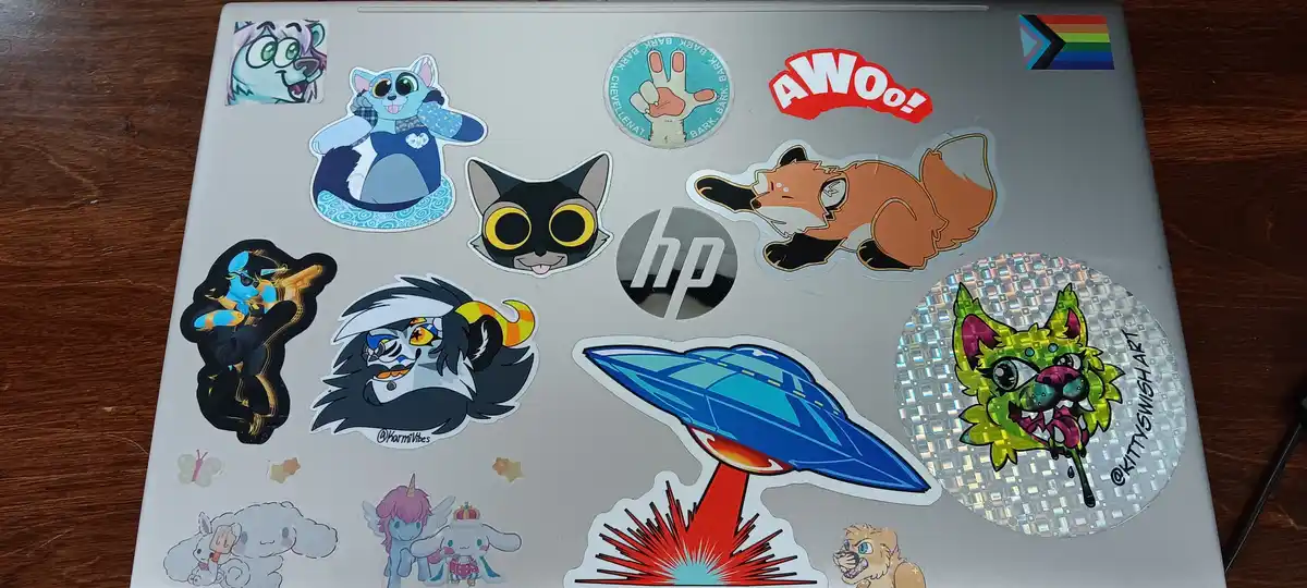 A silver laptop lid is covered in numerous colorful stickers, including cartoon cats, a fox, a UFO, a rainbow flag, and an…
