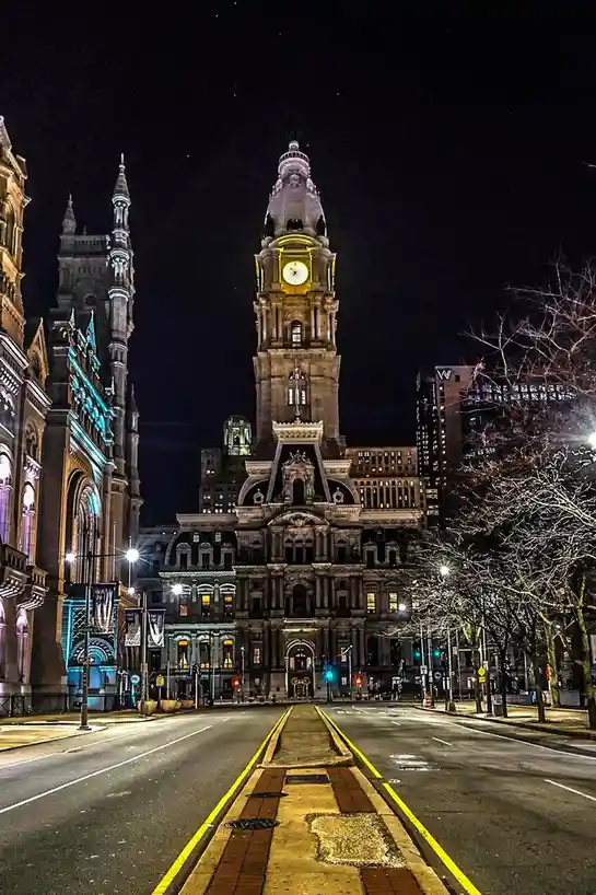 A nighttime photo of Philadelphia City Hall, a grand stone building with a prominent clock tower, viewed from a street with…