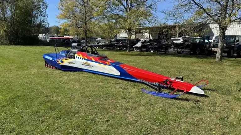a red, white, and blue hydroplane boat with flame decals is lying on its side in a grassy field, surrounded by trees and…