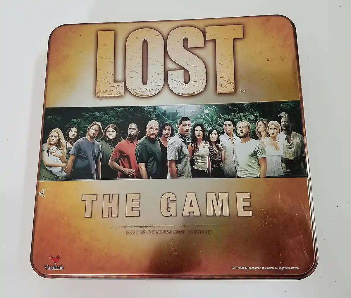 A metal game tin for 'Lost: The Game' shows the TV show's main cast standing in a jungle. The tin has 'LOST' at the top and…