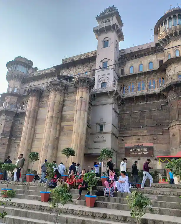 A large, ornate stone building with multiple towers and balconies stands behind wide stone steps where many people are…