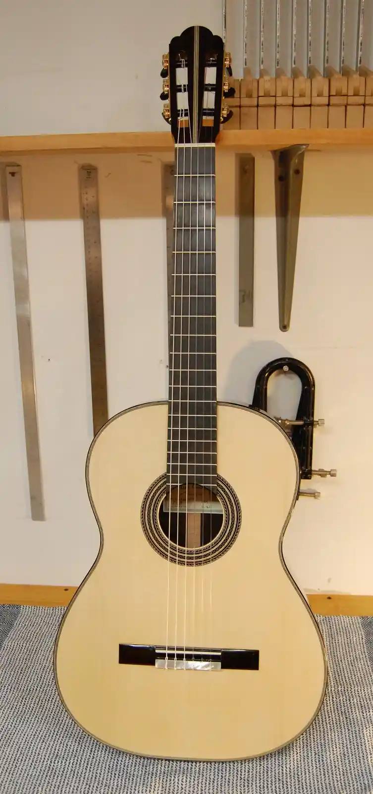 A classical guitar with a black U-shaped lock attached to its body, standing upright against a wall with rulers and tools…