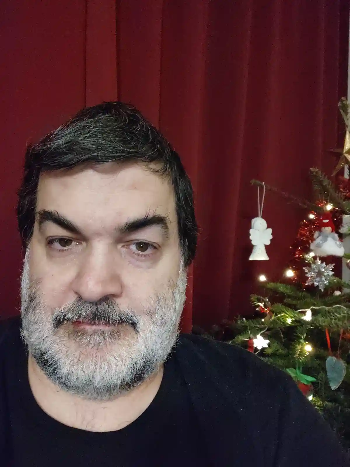 A man with a graying beard and intense stare looks at the camera, standing in front of a red curtain with a decorated…