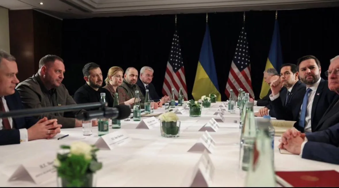 A formal meeting of officials seated at a long table with American and Ukrainian flags behind them. The image is overlaid…