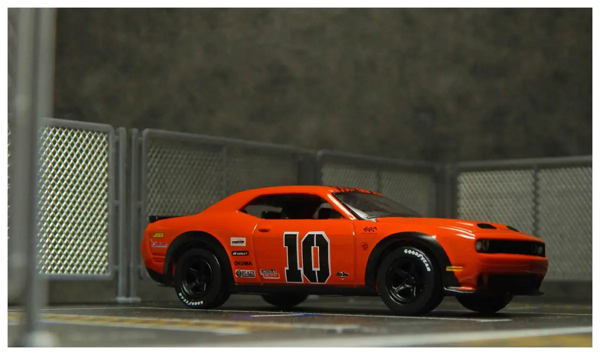 A bright orange die-cast model of a Dodge Challenger race car, number 10, sits on a miniature track with a chain-link fence…