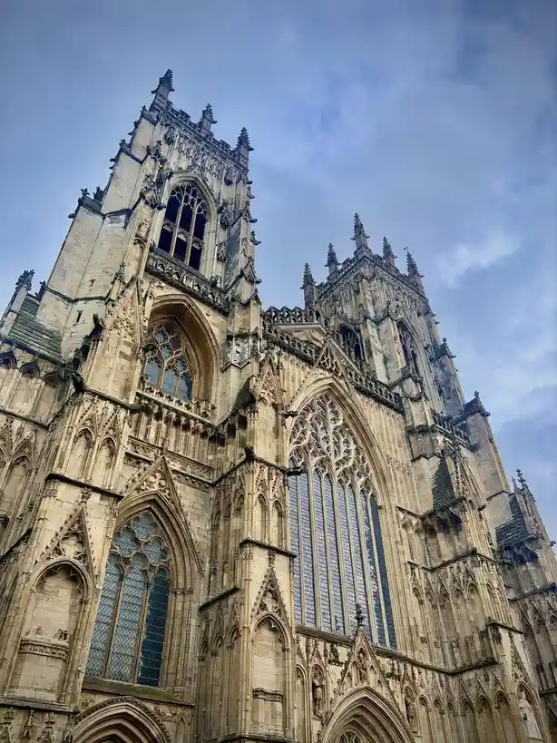 A low-angle photograph shows the ornate, Gothic-style facade of a large stone cathedral with intricate stonework, pointed…