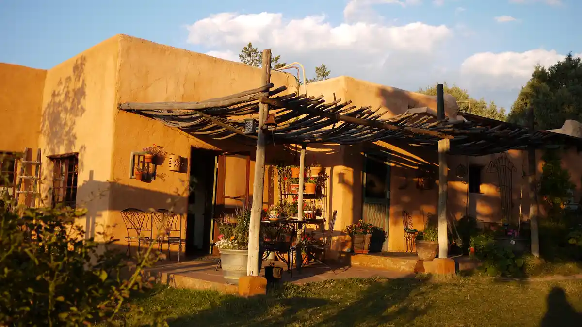 a picturesque adobe-style house with a wooden pergola, potted plants, and outdoor furniture bathed in golden sunset light…