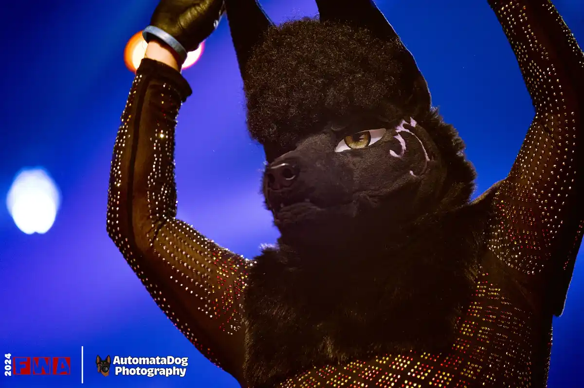 A person in a black furry dog costume with glittery sleeves raises their arms against a blue background. The costume has a…