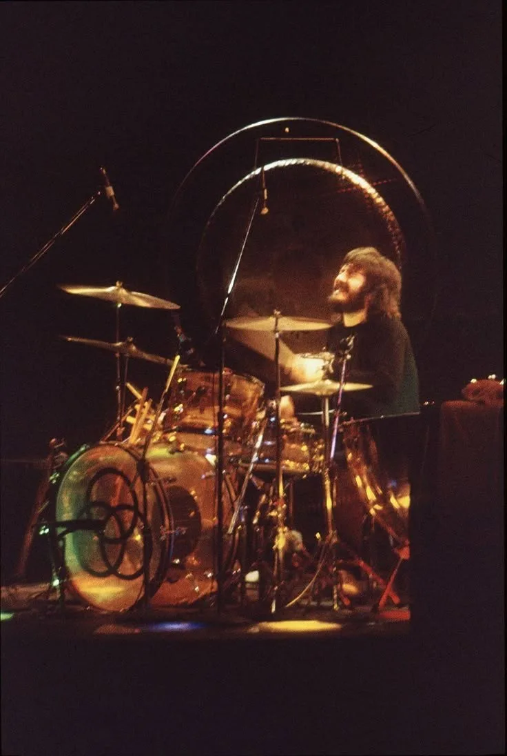 A man with a beard and long hair sits behind a large, ornate drum kit on a dark stage, looking upward with a serene…