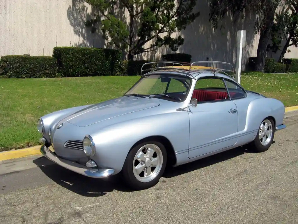 A silver 1960s Karmann Ghia coupe with red interior and chrome wheels is parked on asphalt next to grass and a building. The…