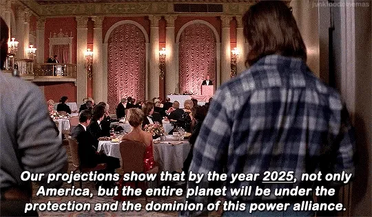 The image shows a scene from the movie 'The Matrix' where a man in a plaid shirt is seated at a formal dinner, listening to…