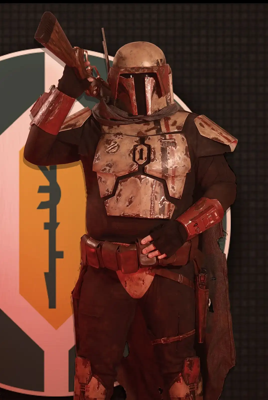 A person in a detailed, weathered Mandalorian armor costume, resembling Boba Fett, holds a blaster rifle over their shoulder…