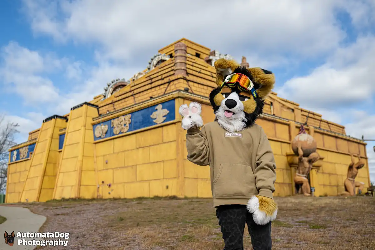 A person in a furry dog costume wearing a beige Supreme hoodie and ski goggles waves at the camera in front of a large…