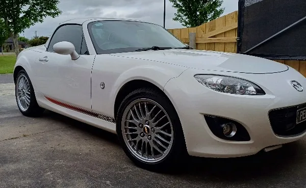A white Mazda MX-5 Miata convertible is parked outdoors on a cloudy day, featuring aftermarket silver multi-spoke wheels and…