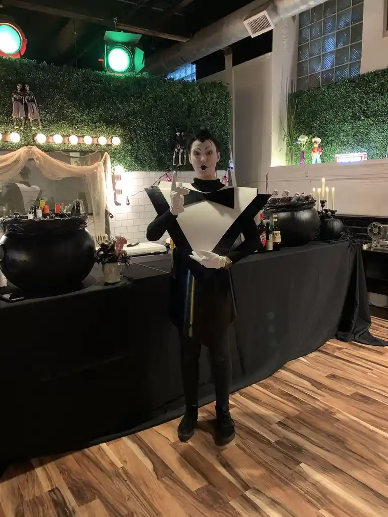 A person in a black and white tuxedo costume with a bowtie and white gloves stands behind a bar decorated for Halloween…