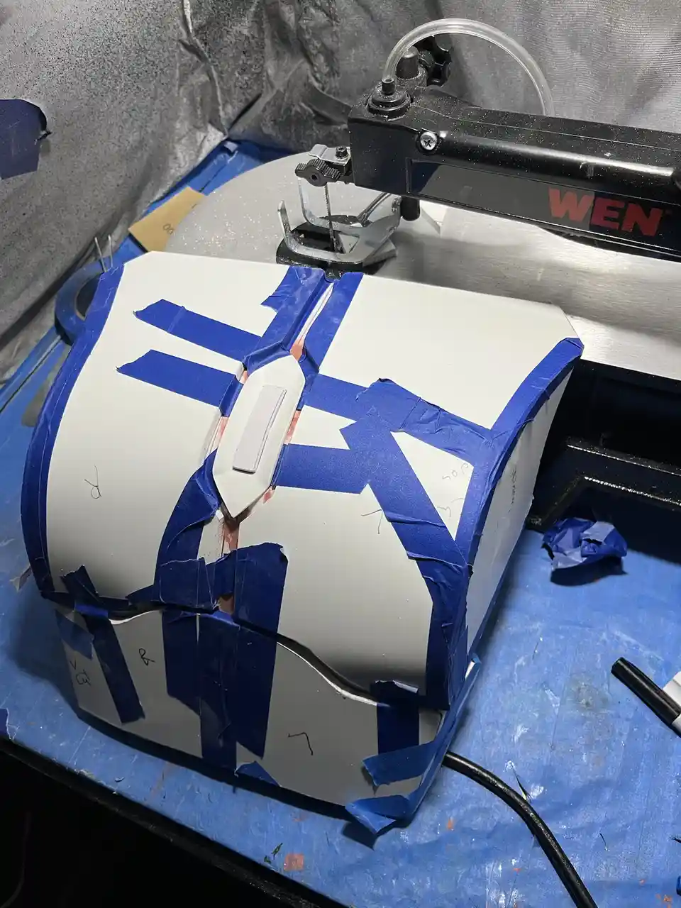 A white, boxy object, possibly a custom-built device or prop, is covered in blue painter's tape with a complex pattern…