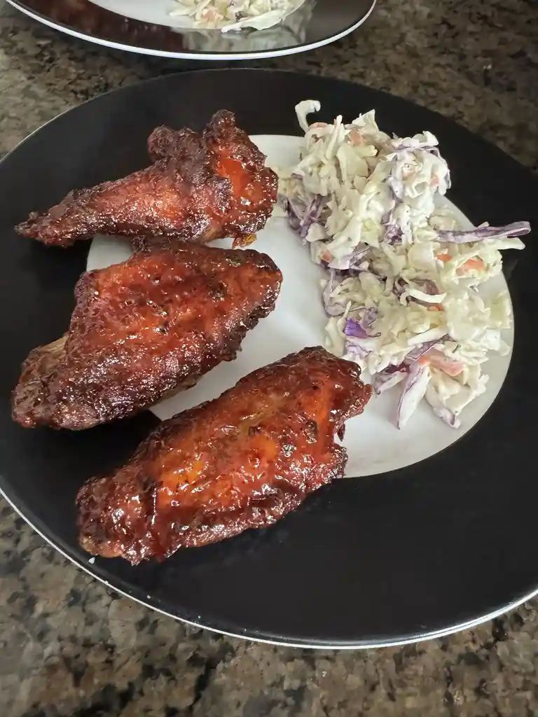 A close-up photo shows a black plate with three glossy, sauce-covered chicken wings and a side of coleslaw on a granite…