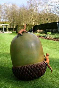 A large, green, artificial acorn sculpture sits on a manicured lawn, with two small, brown squirrel figurines perched on its…