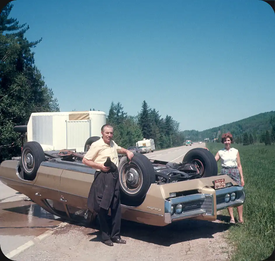 A vintage photo shows a man and woman standing beside a gold Pontiac Catalina that is flipped upside down on a roadside. The…
