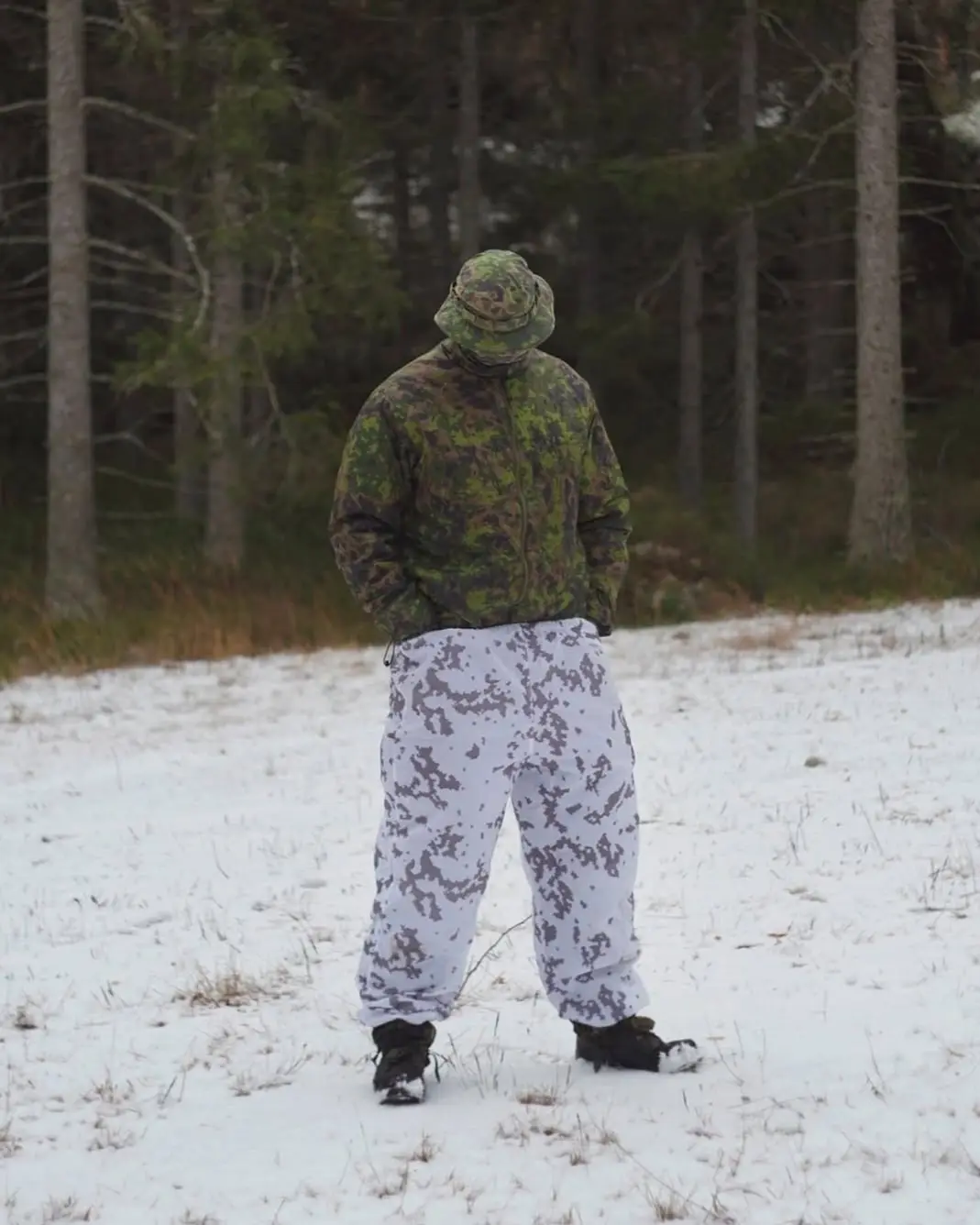 A person stands in a snowy forest wearing a green and brown camouflage jacket and hat, paired with white and grey camouflage…