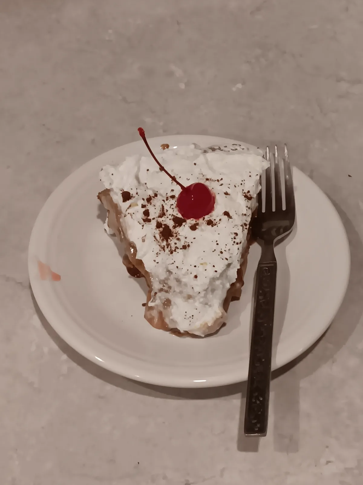 A slice of pie topped with whipped cream, chocolate sprinkles, and a maraschino cherry sits on a white plate with a fork…