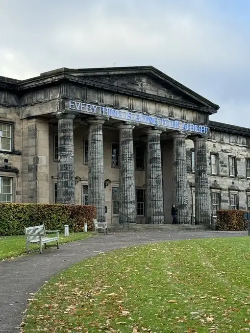 A large, classical-style stone building with prominent columns and a triangular pediment. A blue neon sign is mounted across…