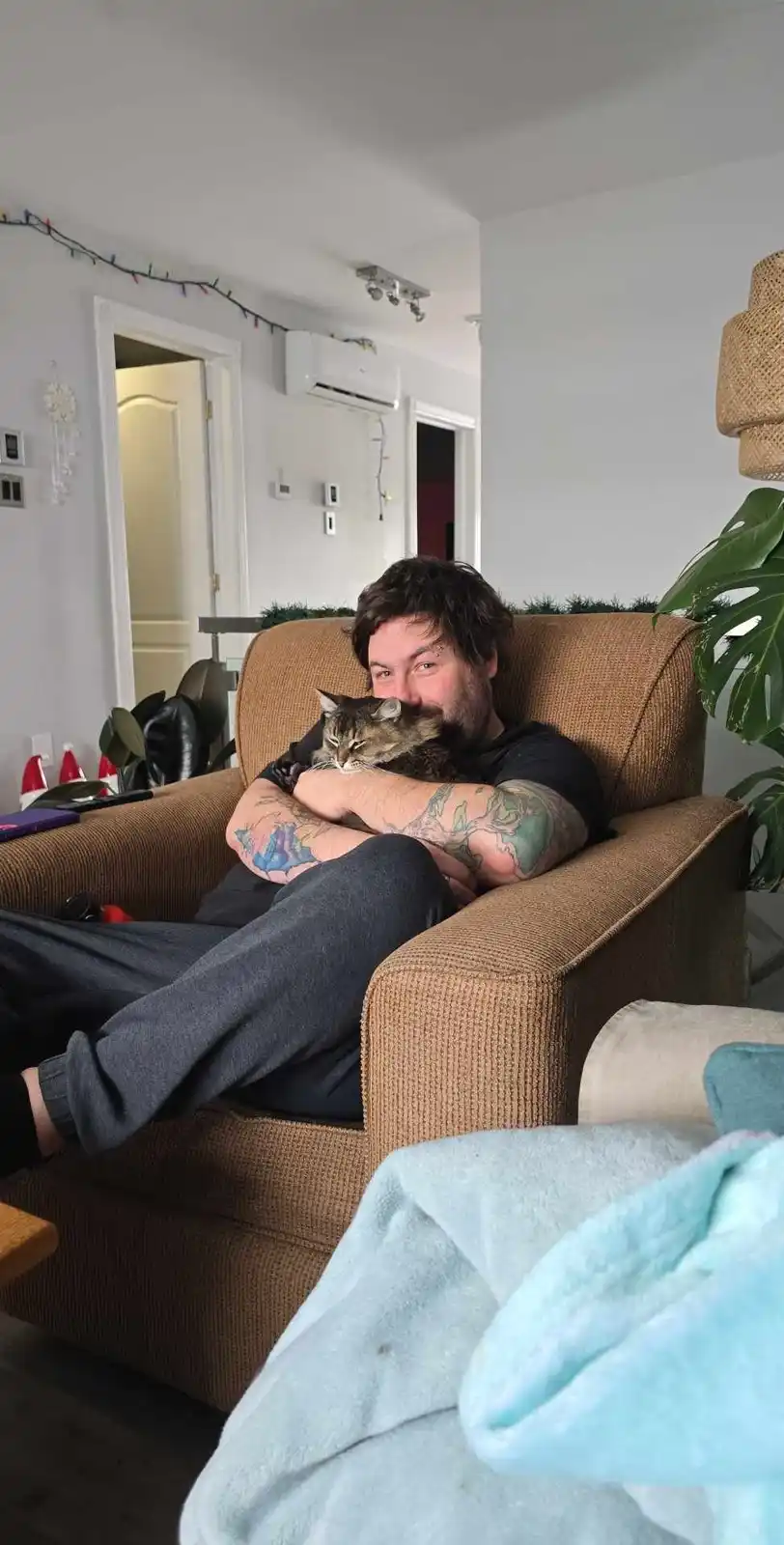 A man with tattoos sits comfortably in a brown armchair, holding a tabby cat close to his chest. He looks directly at the…