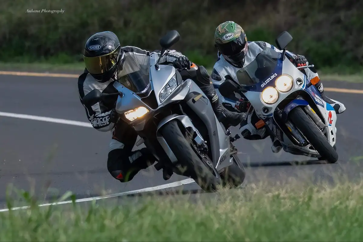 Two motorcyclists race closely on a paved road, leaning into a turn. The rider on the left on a silver Honda wears a black…