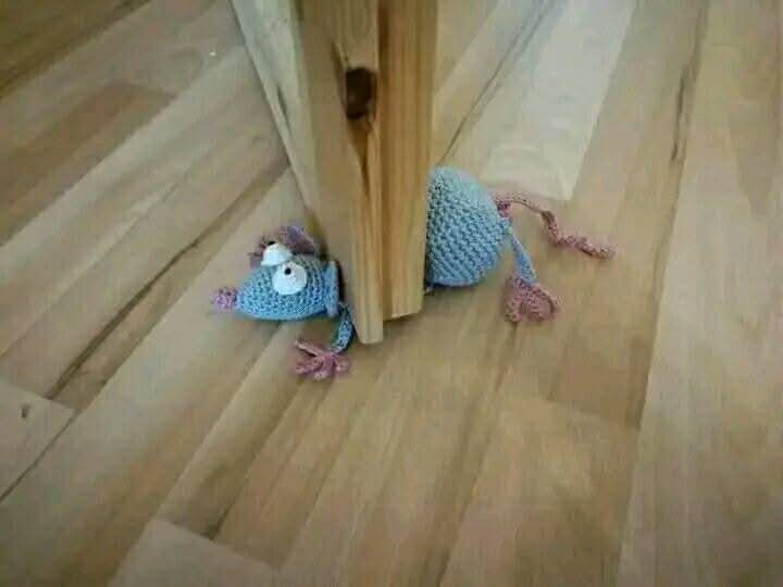 A handmade crocheted mouse toy with large white eyes and pink limbs is peeking out from behind a wooden door frame on a…